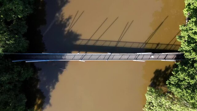 Aerial view of a narrow pedestrian bridge crossing a calm river, surrounded by lush green trees along the banks in Delft, South Holland, Netherlands.