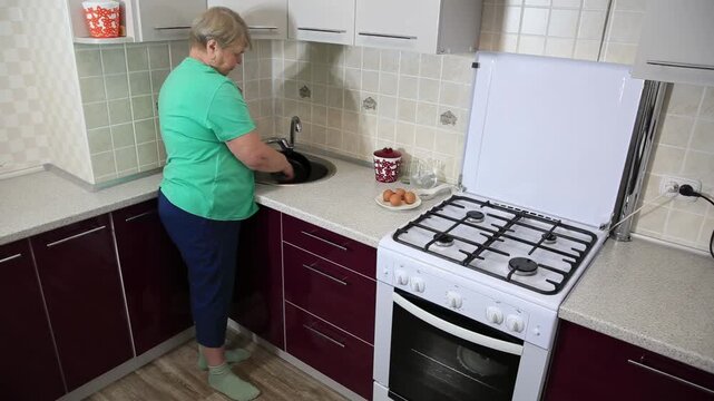 Older woman washing dishes in kitchen sink next to gas stove with eggs on counter, performing daily household chore in modern home environment