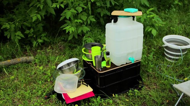 Portable outdoor washing station setup for camping activity, featuring large water container, various cooking utensils, bowls, cups, and soap, surrounded by green grass and foliage.