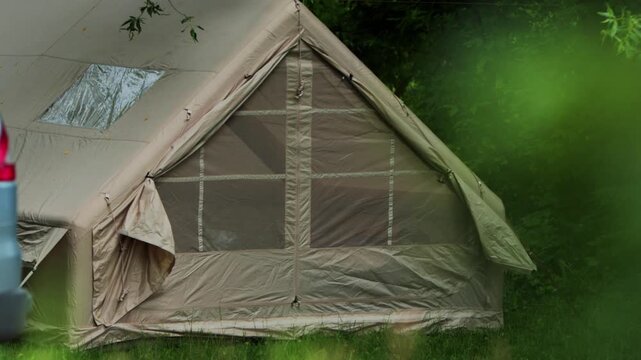 Large beige camping tent with windows and roof vent set up on green grass at edge of forest, suggesting outdoor recreation and temporary shelter in nature