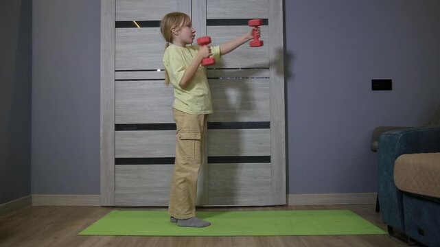 Young caucasian girl performing arm exercises with red dumbbells on green mat indoors. Fitness routine of child for healthy lifestyle at home.