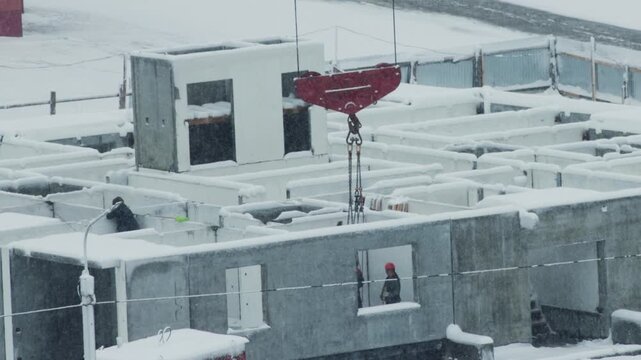 Construction site activity during heavy snowfall, crane moving concrete building section, worker observing progress amid winter conditions