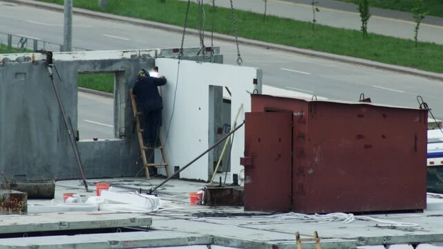 Welder On Ladder Joins Concrete Panels. Industrial Worker Uses Welding Machine On Site.