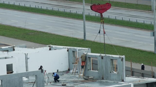 Construction workers assemble pre-fabricated concrete wall panels on building site using large red crane hook overhead by busy road with cars