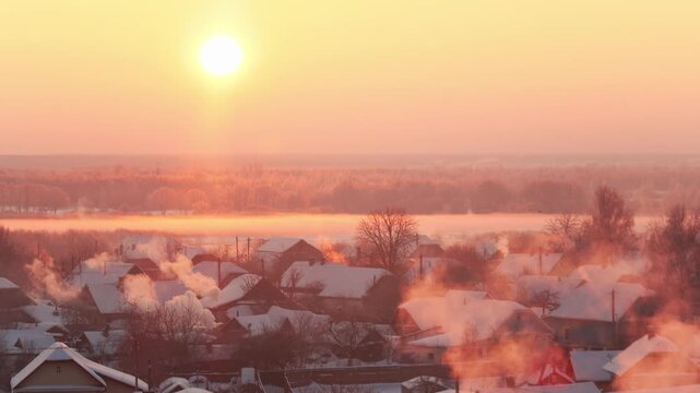 Golden sunrise over frozen winter village with smoke plumes rising from chimneys. Frost-covered trees and misty river create serene morning landscape. Copy space for text