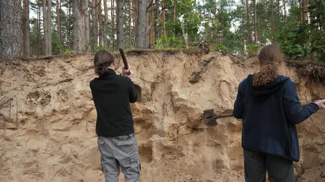 Two white teens sampling sand cliff at forest edge with shovels, methodical strikes and careful collection of soil samples, observational fieldwork and measurement, educational research atmosphere