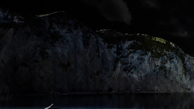 A rocky cliff is seen at night. The top of the cliff has some light. Water reflects the dark surroundings. Clouds are present in the sky, creating an evening scene.
