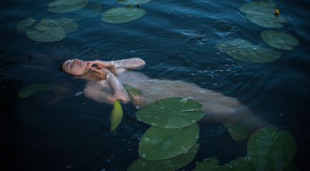 Ethereal woman floating in dark lake water among green lily pads, mystical water nymph or goddess in white shirt, fine art fantasy concept © T.Den_Team