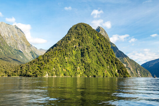 View of Mitre Peak rising above the calm waters of Milford Sound, surrounded by lush green rainforest and steep cliffs under a clear blue sky in Southland Region, New Zealand.