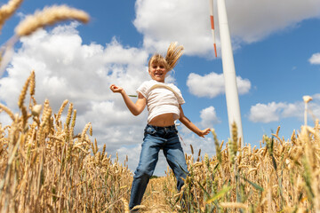Fototapeta premium Laughing girl runs through golden wheat field with open arms, wind turbine rising behind under blue sky. Sunny scene suggests childhood joy, clean energy, harvest, nature, freedom, sustainability
