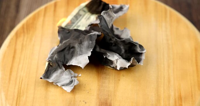 Horizontal view of twenty dollar cash money bills burning slowly while rotating on a wooden platform over a dark table workspace
