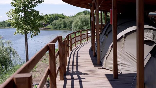 A row of modern camping tents stands under a protective wooden pavilion on a boardwalk next to a calm lake. Concept of luxury camping, summer vacation, and peaceful outdoor recreation.
