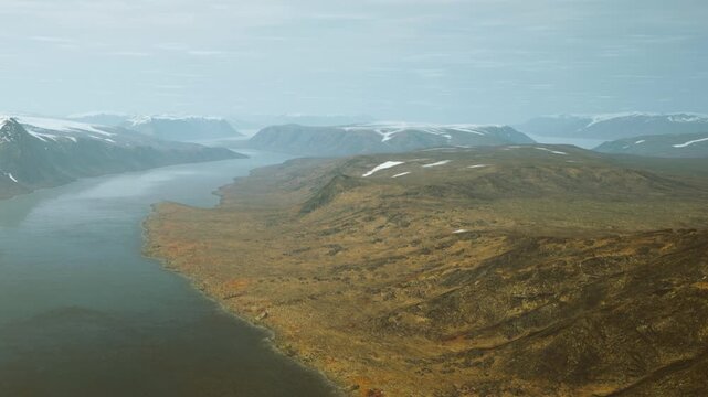 A large river flows through a valley surrounded by mountains. Snow capped peaks rise in the distance. The terrain is mostly brown and gray with some patches of green.