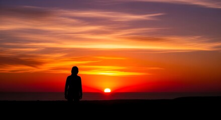 Fototapeta premium Silhouette of a person standing on a dark shore watching a vivid golden sunset across the tranquil ocean waves under a dramatic summer sky.