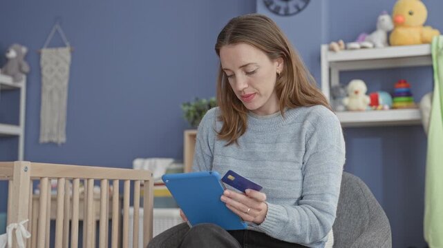 Woman holds a tablet and creditcard while tapping the screen, seated beside a wooden crib in a blue nursery with stuffed toys on shelves; planning.