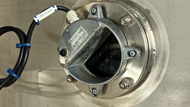 Close-up of a stainless industrial inspection window with bolted flange, sensor cable and viewing port on clean processing equipment.