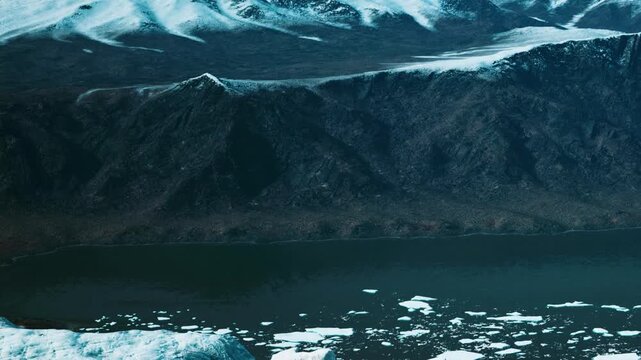 Ice and snow cover the mountains while pieces of ice float in the water. The sun sets in the background, creating a transition from day to night along the landscape.