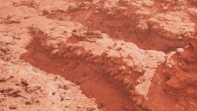 Red rock formations are visible on a dry terrain. The sunlight casts shadows on the rocks, highlighting their shapes and textures. The ground appears cracked and dusty.