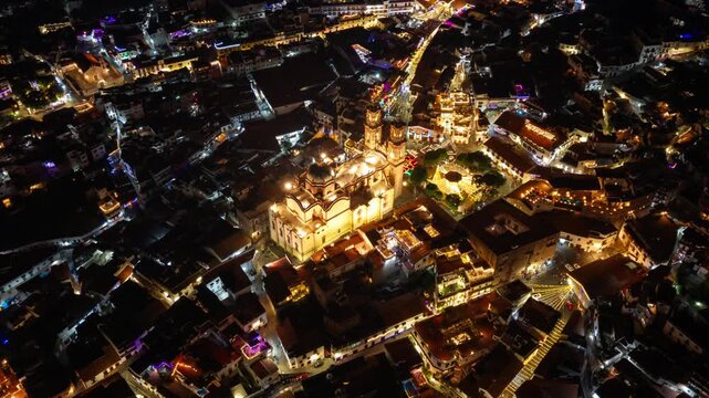 Tilt-down aerial hyperlapse of the Parish Church of Santa Prisca and San Sebastian at night in Taxco, Mexico, featuring lights and car trails, colonial architecture
