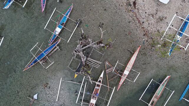 Straight down overhead aerial drone shot of colourful outrigger bangka boats beached at low tide on a rocky mudflat at Punta Bilar Surigao City Philippines with a driftwood stump at centre.