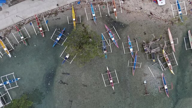 High overhead aerial drone view of colourful traditional outrigger bangka boats clustered in a shallow bay cove at Punta Bilar Surigao City Philippines with a seawall visible at the top of frame.