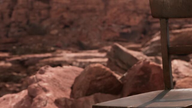 A wood chair is set among rocks in a canyon area. The landscape shows rugged stone formations and a dry environment. Sunlight highlights the textures of the rocks.