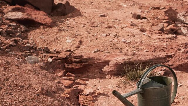 A watering can sits on the dry ground surrounded by rocks and sparse grass. The sun shines brightly in the clear sky. The area appears to be remote and arid.