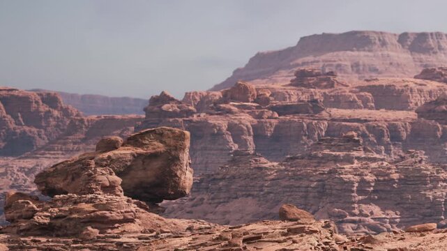 Rocky mountains rise in the distance under a clear sky. The landscape is dry with scattered stones. Sunlight highlights the textures of the rocks. Nature is vast and open.