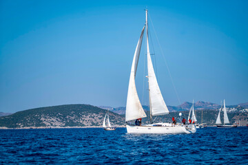 Fototapeta premium Luxury sailing yachts and white boats cruising on the deep blue Mediterranean sea during an active summer regatta ocean vacation with a scenic coastal resort town and mountains in the background