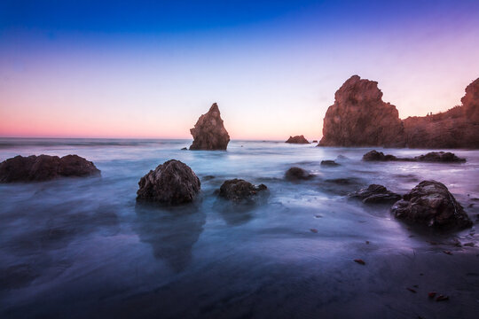View of rocky sea stacks on a sandy beach at sunset with calm waves and soft light in Malibu, California, United States.