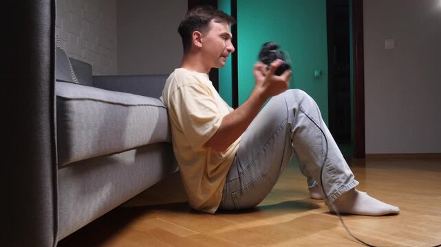 Young man sitting on the floor in a living room, holding a controller and cheering with joy and excitement after winning a video game, enjoying his hobby