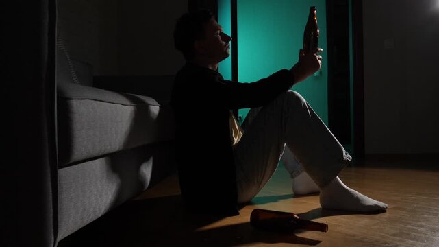 Young man sitting on the floor in a dark room with a bottle of beer, struggling with loneliness and mental health issues, representing alcoholism, depression, and self destruction