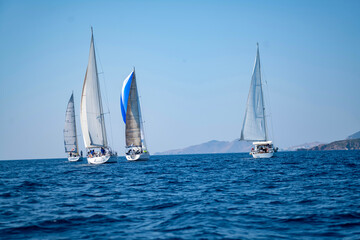 Fototapeta premium Luxury sailing yachts and white boats cruising on the deep blue Mediterranean sea during an active summer regatta ocean vacation with a scenic coastal resort town and mountains in the background
