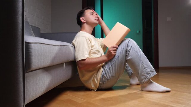 Young man sitting on the floor leaning against a sofa in a modern apartment, holding a book and covering his face while deep in thought