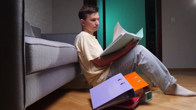Man sitting on the floor leaning against a sofa, holding an open folder and studying documents, surrounded by colorful binders, working from a home office environment