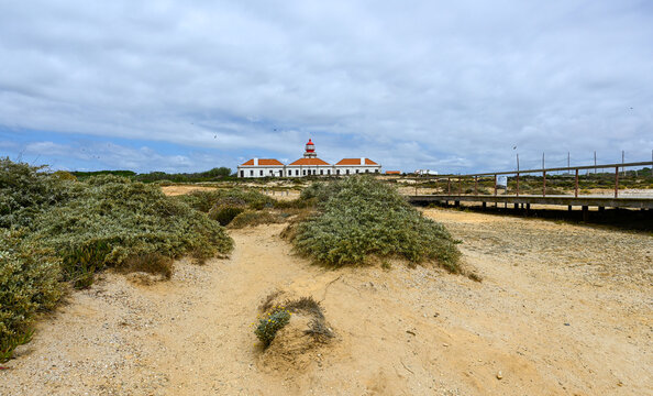 View of a historic lighthouse with a red lantern room situated on a sandy coastal landscape with low-lying shrubs and a wooden walkway in Algarvia, Azores, Portugal.