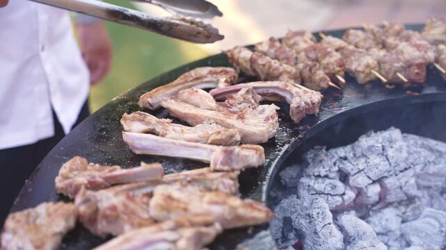 People are cooking meat on a grill over charcoal in an outdoor setting. The sun shines down as they prepare food for a gathering in the afternoon.