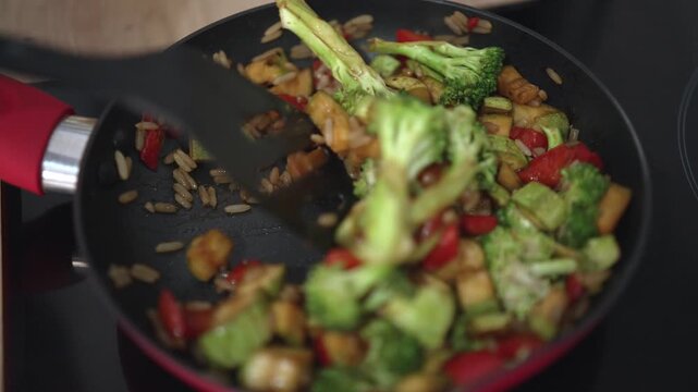 Sliced bell peppers and tofu are cooked with broccoli and onions in a frying pan. The heat creates steam as the ingredients mix together in a home kitchen setting.