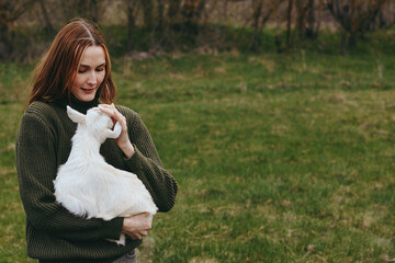 Fototapeta premium Casual woman outdoors cradles a white goat kid, smiling softly in a green field. The scene conveys a gentle lifestyle and close bond with animals, capturing calm rural moments.