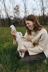 Fototapeta premium A woman in a knitted sweater bottle feeds a young goat outdoors, amid grass and trees. A tender moment of caregiving, farm lifestyle, and animal bonding in a natural setting today.