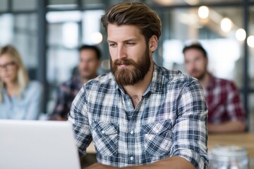 Obraz premium Focused bearded man in plaid shirt working on laptop in modern office with blurred colleagues in background