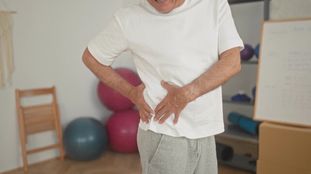 Man clutching hip and lower back, grimacing while holding side amid exercise balls and whiteboard in studio; pain recovery.