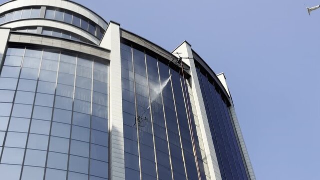 Drone is washing windows. Drone spraying water from a suspended nozzle onto upper windows of a modern building. Red hose hangs below the aircraft against blue sky, showcasing facade cleaning