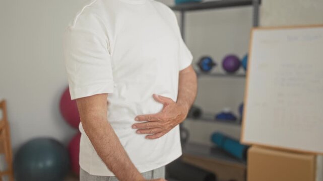 Man pulls purple resistance band with right hand while holding abdomen in exercise studio; rehab recovery determination.