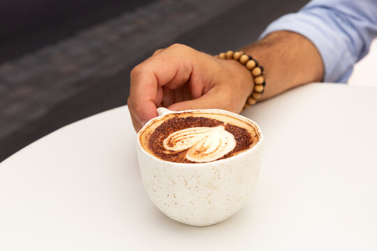 Caf&eacute; cappuccino en gros plan dans une tasse avec un latte art coeur, pos&eacute; sur une table blanche de terrasse de caf&eacute;, bras et main d'un homme avec bracelet bois et manche de chemise bleue retrouss&eacute;e