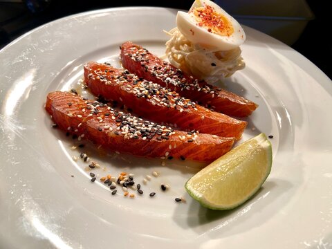 Close-up of smoked salmon slices served with a soft-boiled egg, lime wedge, and sesame seed garnish on a white plate. Fresh seafood dish ideal for brunch, breakfast, healthy eating, gourmet dining, pe