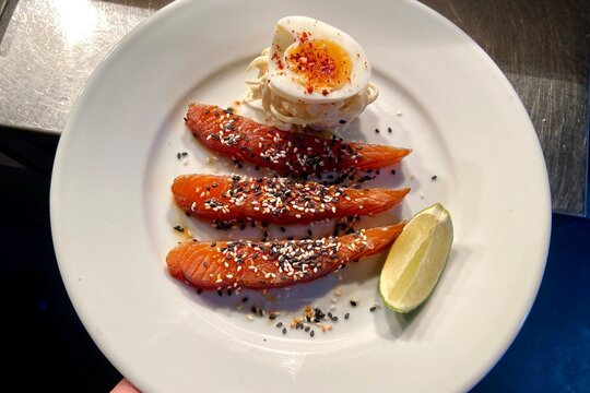 Close-up of smoked salmon slices served with a soft-boiled egg, lime wedge, and sesame seed garnish on a white plate. Fresh seafood dish ideal for brunch, breakfast, healthy eating, gourmet dining, 