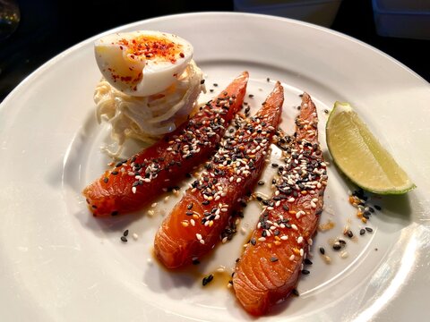 Close-up of smoked salmon slices served with a soft-boiled egg, lime wedge, and sesame seed garnish on a white plate. Fresh seafood dish ideal for brunch, breakfast, healthy eating, gourmet dining, pe