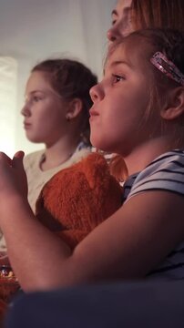 Two girls are watching something intently, showing expressions of focus. The girl on the right holds a stuffed toy while both girls sit close together.