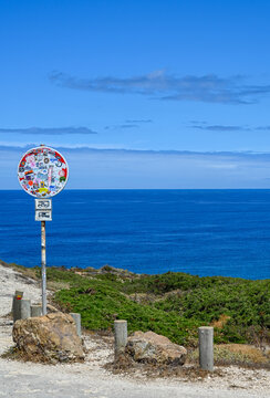 View of a road sign covered in colorful stickers standing on a coastal path overlooking the deep blue Atlantic Ocean with green shrubs and rocky terrain in Algarvia, Azores, Portugal.